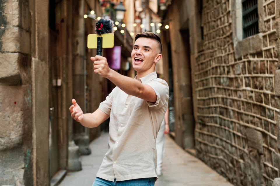 A young man in casual attire films himself with a handheld camera in an alleyway, appearing enthusiastic and animated