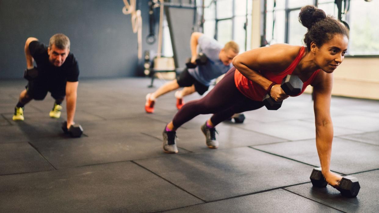  in the foreground, we see a woman wearing atheletic clothing and smiling as she lifts a dumbell off the ground while holding a plank position. Two men are completing the same exercise in the background 