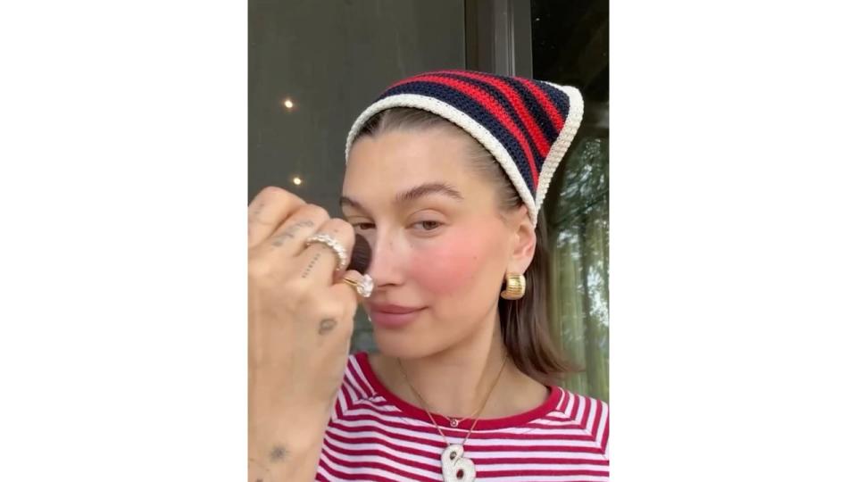 Hailey Bieber does her make up wearing a stripe t-shirt and matching crochet bandana