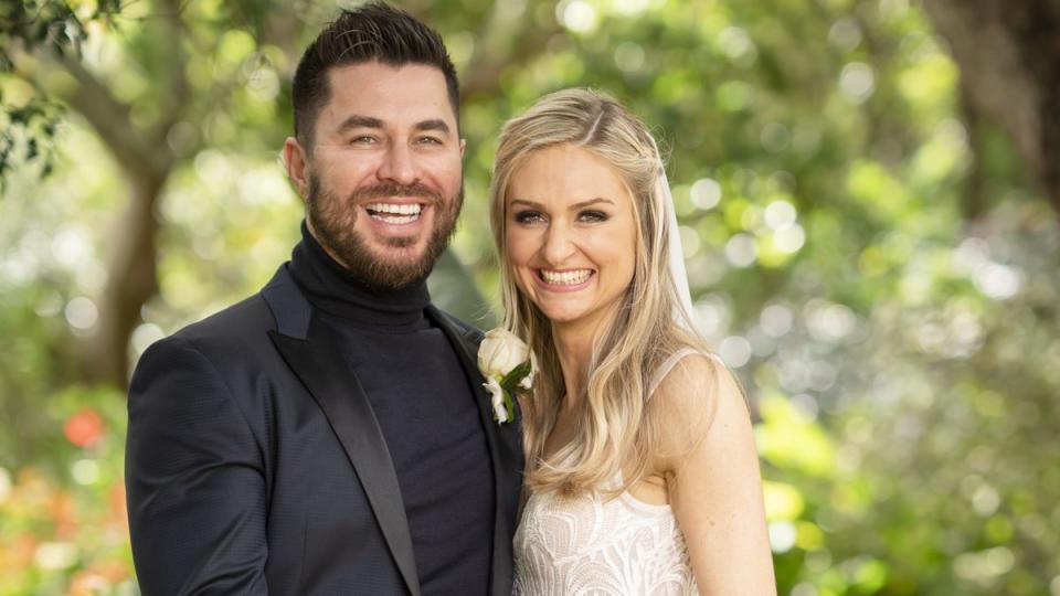 2021 MAFS couple Joanne and James in a garden at their show wedding. He wears a dark turtle-neck and dark suit with satin lapels and a white rose. She wears a lace gown and her blonde hair is loose..