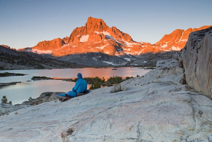 A lone backpacker with a blue stocking cap sits on a slab of granite rock and watches the early sun beams of the morning light hit Banner Peak behind Thousand Island Lake in the Ansel Adams Wilderness.