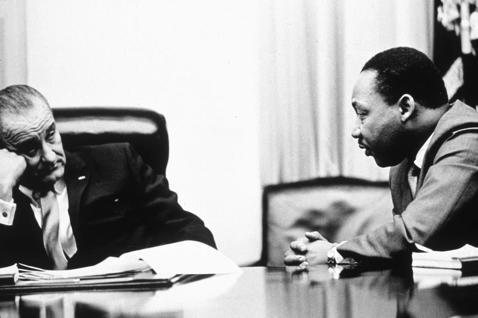 A  middle-aged white man dressed in a business suit sits on a leather chair and listens to a black man sitting next to him.