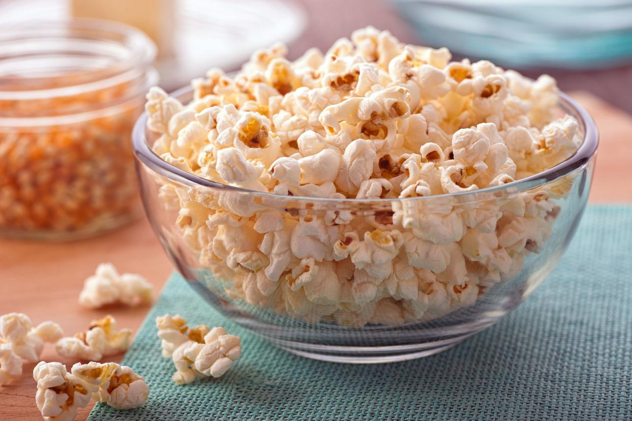 Glass bowl of popcorn with a few scattered popcorn pieces on a blue napkin with a blurred background of popcorn kernels and glassware