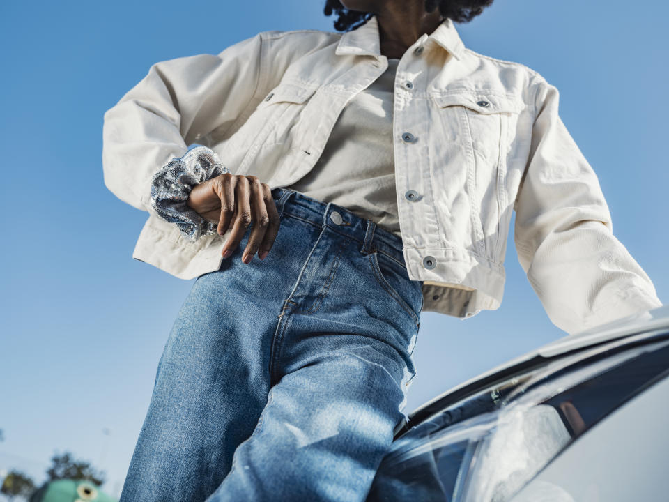 Person in casual attire with hand on hip standing by a car, clear sky in the background