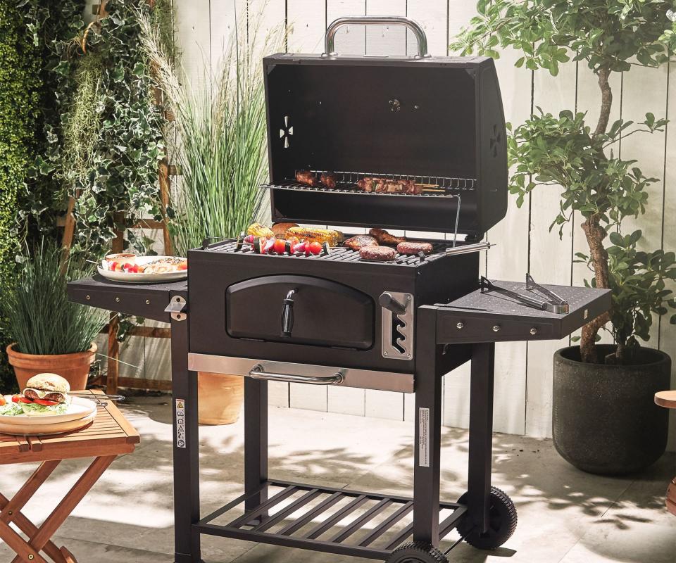 A black charcoal gril in a back garden, against a white fence, frying food