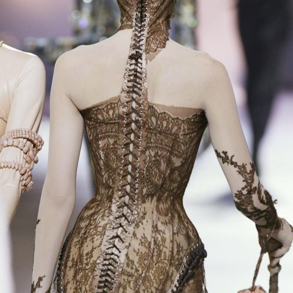 Model in a fitted, lace-detail bodysuit with a spine-like adornment down the back, on a fashion runway