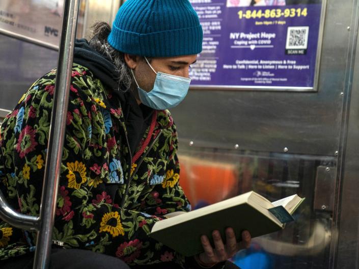 A man wearing a blue cap, face mask and colorful jacket reads a book as he commutes on the New York City subway.