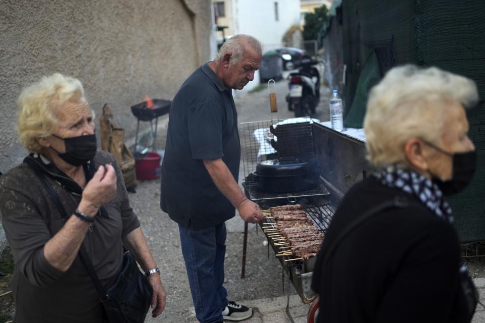 A man grills pork skewers or souvlakia in Greek, as two women arrive for the church service at an Orthodox temple in Hasia, northwestern Athens, Greece, Sunday, Aug. 14, 2022. The Dormition of the Virgin Mary (or Mother of God as the Greeks usually refer to her) is celebrated on Aug. 15. The religious event is coupled with midsummer festivities, known as Panigiria, that often last more than a day with music, culinary feasts and, in many cases, flea markets. (AP Photo/Thanassis Stavrakis)