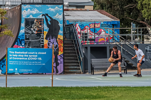 Two men not observing social distancing playing basketball in Prahran with a sign outside the court reading that the court is closed.