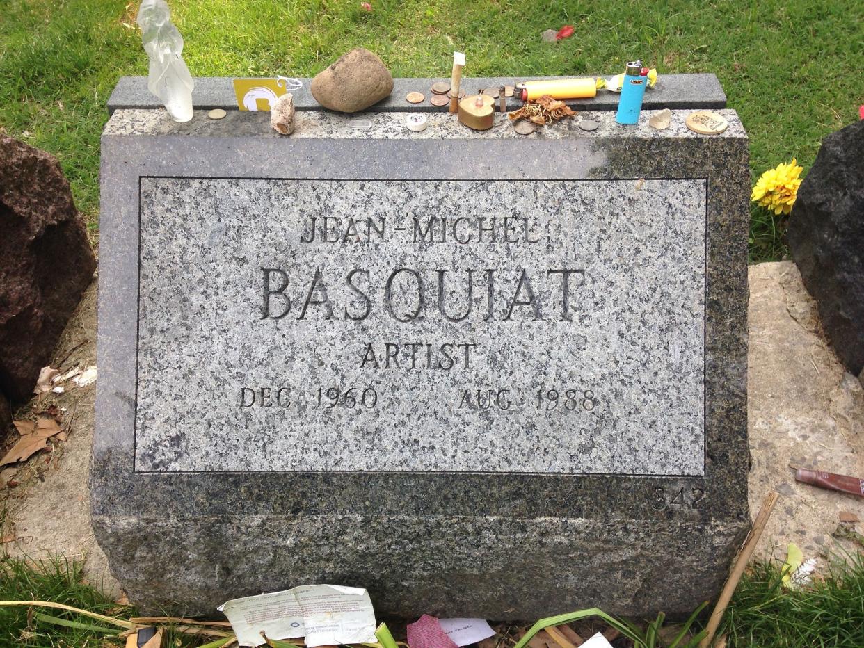 Closeup of Jean-Michel Basquiat's grave in the Green-Wood Cemetery, Brooklyn, New York with several crystals, coins, and other keepsake items on top of it with two large rocks on both sides and grass in the background