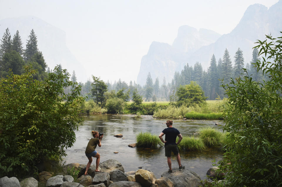 German tourists Stephanie Schultz, left, photographs Kai Rudolph, right, along the Merced River in Yosemite Valley as smoke from the Ferguson Fire hangs in the air Tuesday, July 24, 2018, in Yosemite National Park, Calif. The heart of Yosemite National Park, where throngs of tourists are awe-struck by cascading waterfalls and towering granite features like El Capitan and Half Dome, will be closed as firefighters try to corral a huge wildfire just to the west that has cast a smoky pall and threatened the park's forest, officials said Tuesday. The closure is expected to last through Sunday. (Eric Paul Zamora/The Fresno Bee via AP)