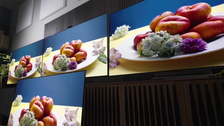 A platter of flowers and fruit against a blue background compared on a Samsung S95C, Sony A95L, LG G3, and Sonly A95K TV. 