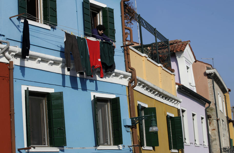 In this image taken on Thursday, Jan. 16, 2020, an elderly woman hangs laundry outside a window, at the Burano island, Italy. The Venetian island of Burano's legacy as a fishing village remains the source of its charms: the small colorful fishermen's cottages, traditional butter cookies that were the fishermen's sustenance at sea and delicate lace still stitched by women in their homes. As the island's population dwindles, echoing that of Venice itself, so too are the numbers of skilled artisans and tradespeople who have kept the traditions and economy alive. (AP Photo/Luca Bruno)