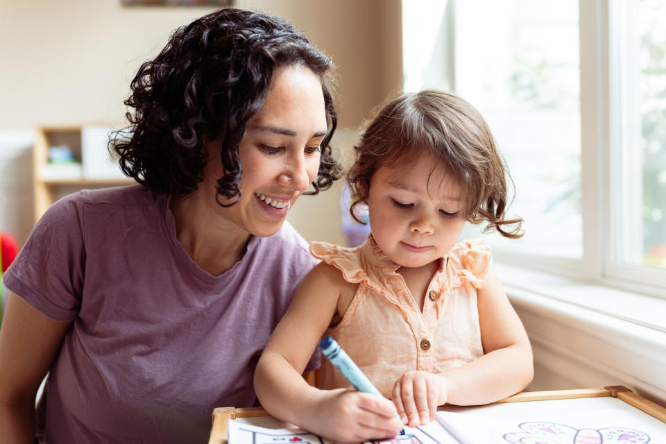 An Eurasian woman who is applying Montessori education techniques while homeschooling her preschool age daughter smiles encouragingly as the young girl colors with a crayon.