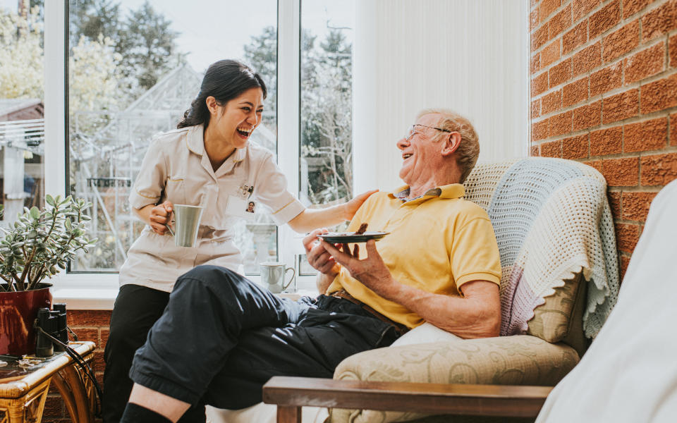 A friendly, young, female carer visits an elderly male in his home. She has made them both a cup of tea and prepared him a snack. She perches beside him as he eats some cake and chats with her. They are comfortable with each other as she reaches in to touch him on the shoulder and they share a joke together.