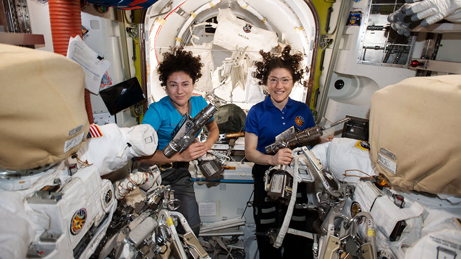 two woman float behind spacesuits in the international space station. an open module door is visible behind