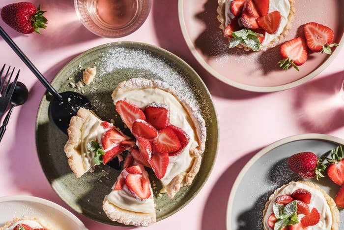 Plates of strawberry-topped tarts with cream and mint leaves are arranged on a pink table, accompanied by a glass of pink beverage