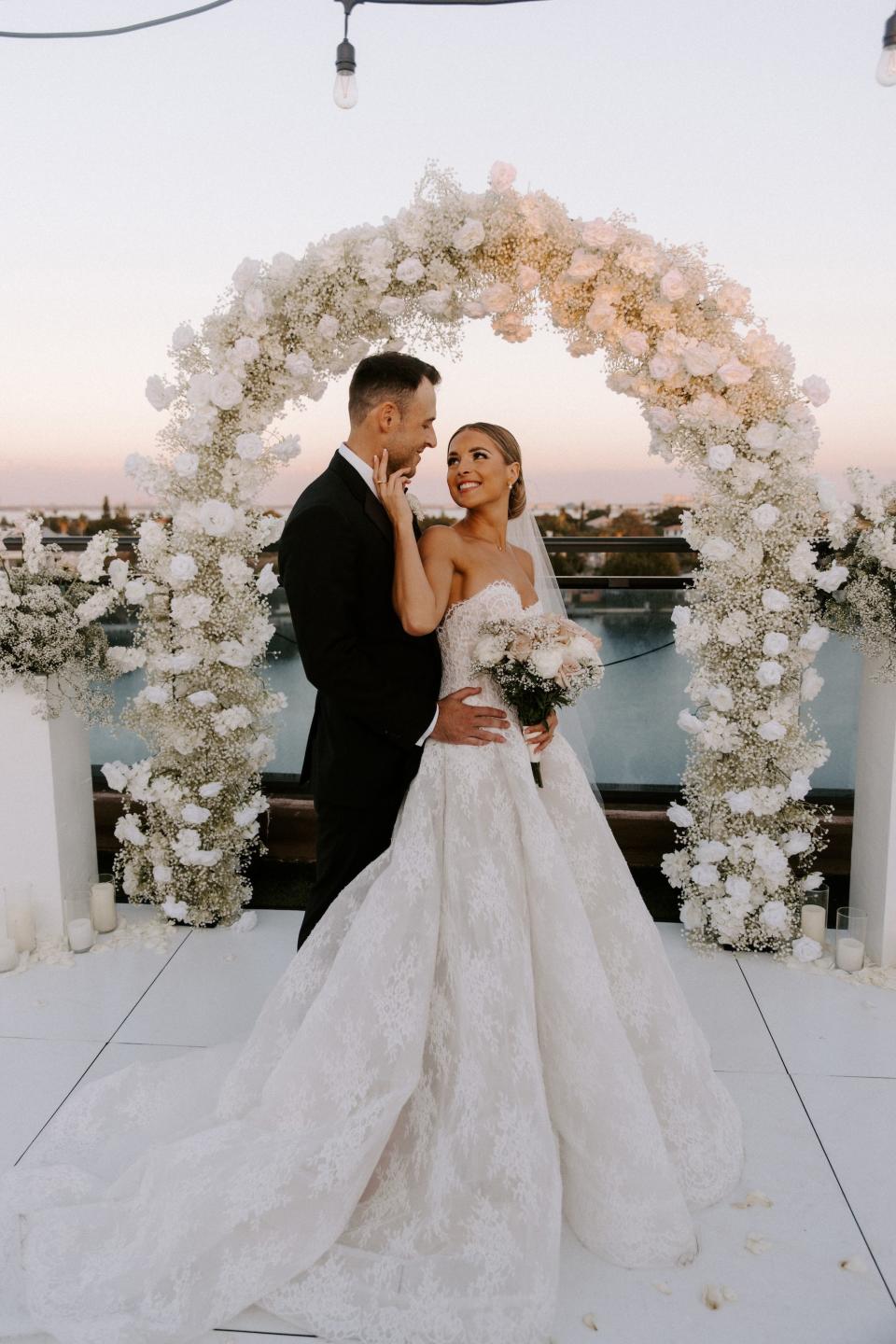 A bride embraces her groom's face in front of a floral arch. 