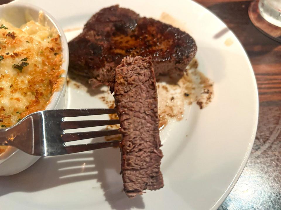 A piece of steak on a fork. The rest of the steak is on a white plate with a small bowl of mac and cheese.