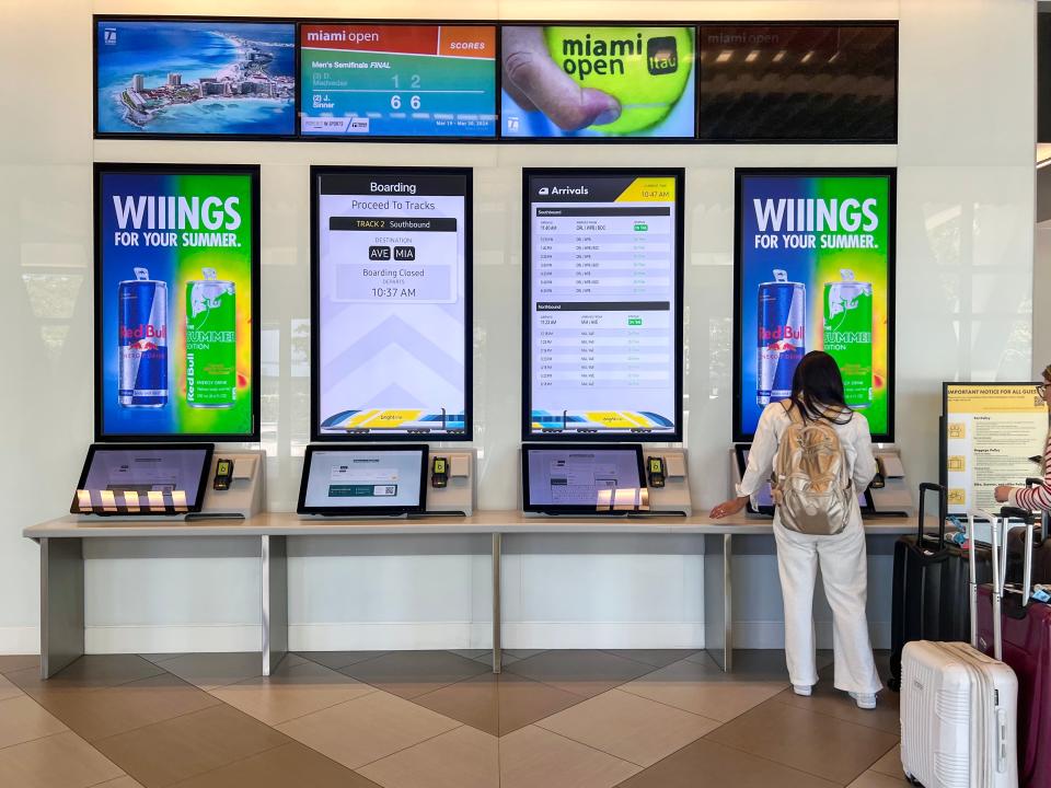 A series of monitors show arrival and departure times for Brightline trains, as well as an ad for Red Bull.  A woman in a cream outfit wearing a gold backpack stands at a kiosk with black and white luggage nearby.