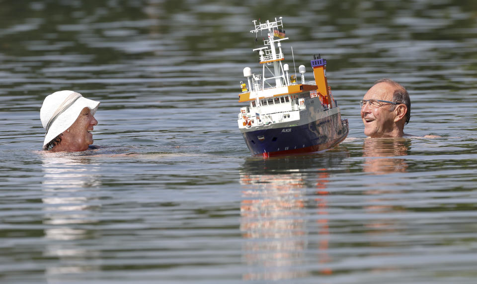 A couple swims beside a model making boat in a lake in Ertingen, Germany, Wednesday, June 26, 2019. A heatwave hits Germany and Europe with temperatures near 40 degrees Celsius. (Thomas Warnack/dpa via AP)