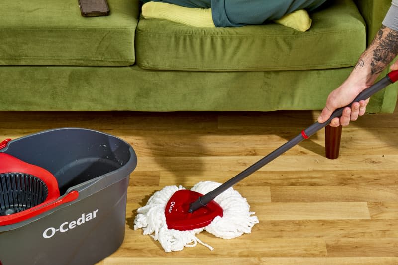angled shot of someone mopping a light wood floor, with an O-cedar mop and bucket.