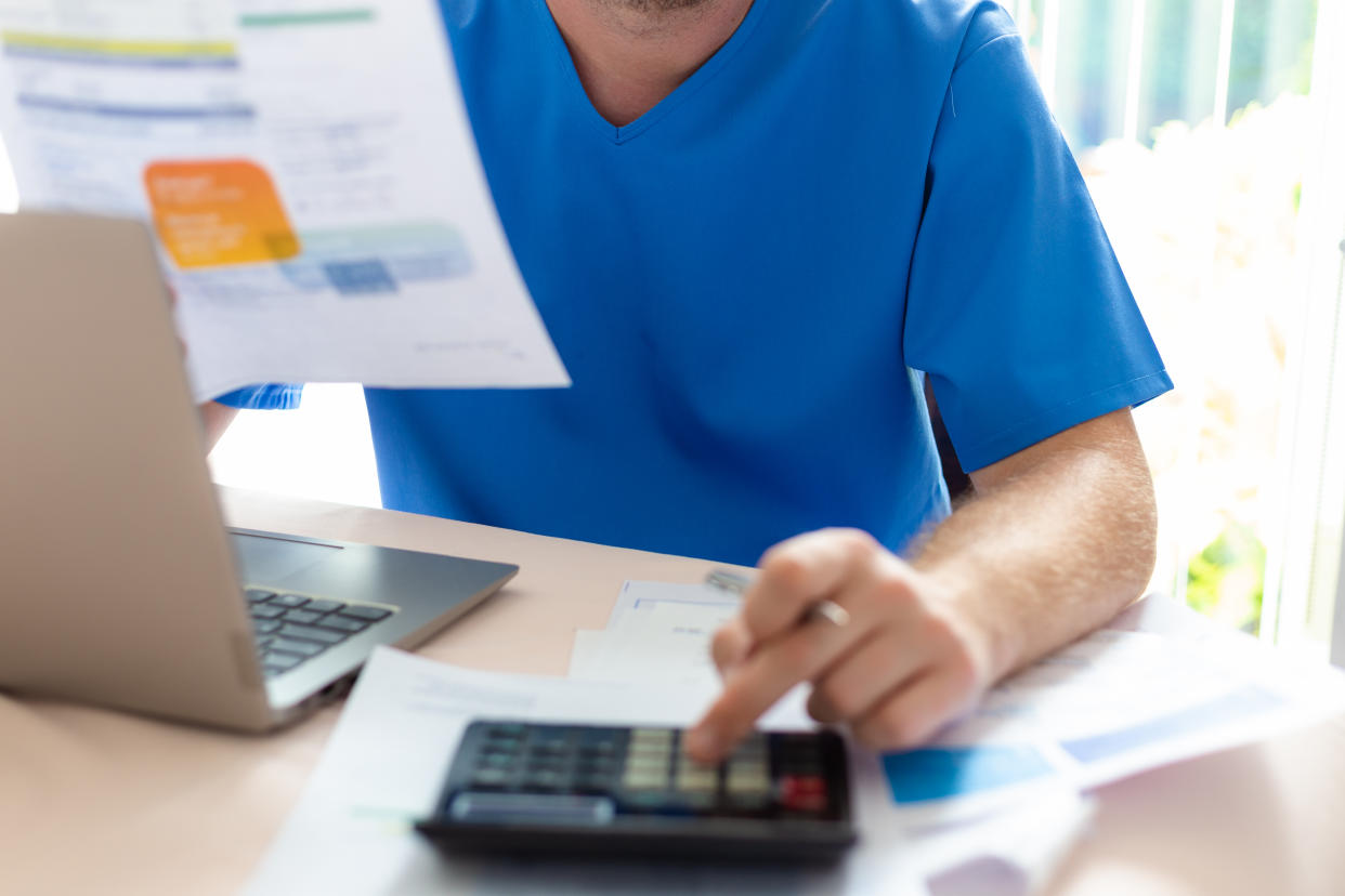 A male nurse in his 30s checks his energy bills at home. He is still wearing scrubs as he has just come home from work. The man has a worried expression as he examines the paper documents.