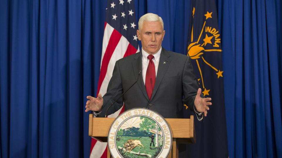 mike pence stands behind a podium featuring a state of indiana seal, he speaks and extends his hands out in front of him, he is wearing a gray suit jacket with a white shirt and red tie, behind him are blue drapes, an american flag, and an indiana state flag