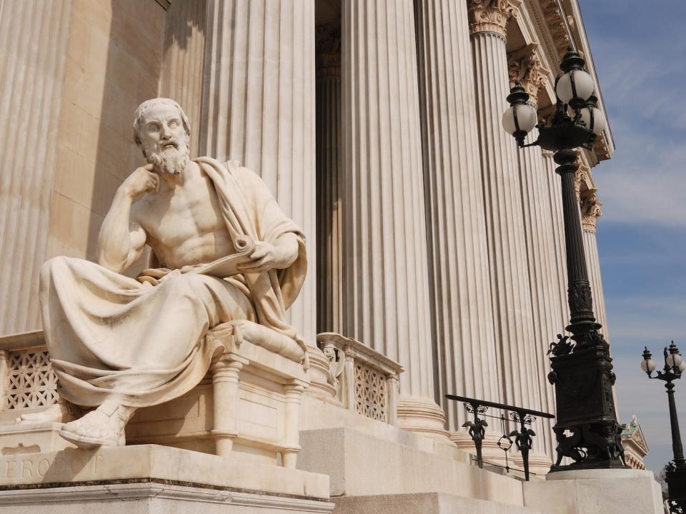 A statue shows a roman man in a toga sitting and leaning in. The pedestal reads "Herodotus"