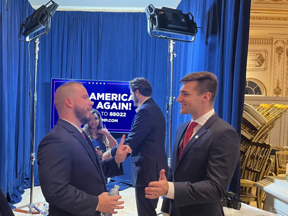 Two men in suits stand next to blue curtain