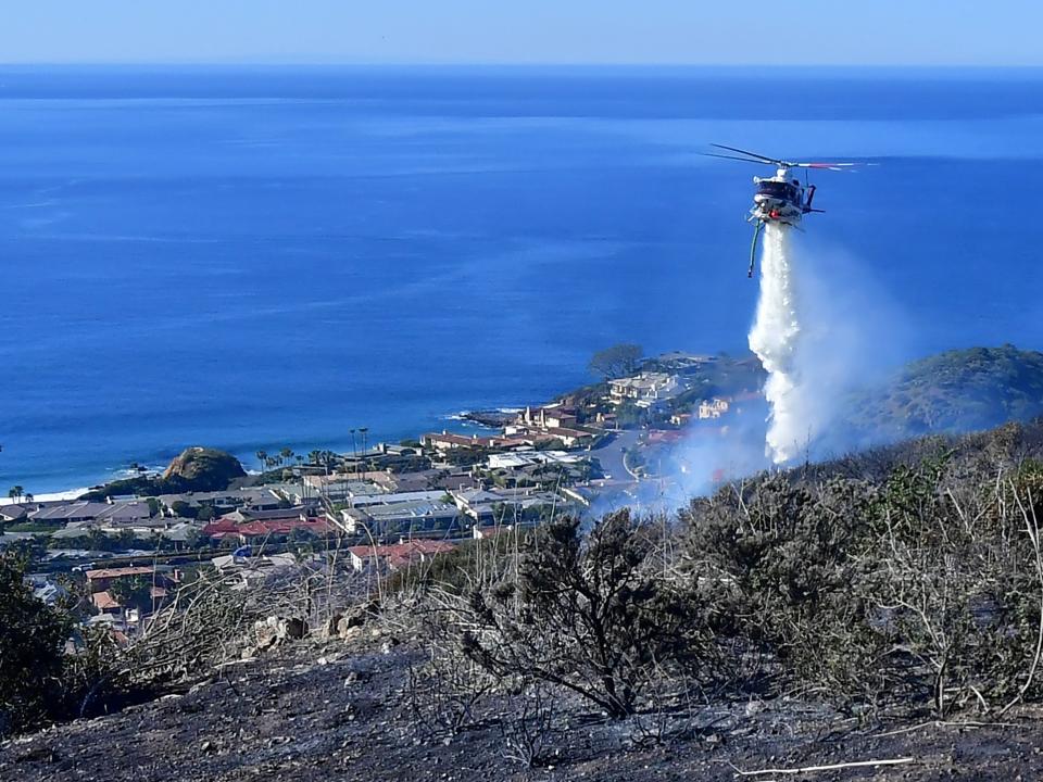 A helicopter drops water on a hillside overlooking expensive homes.