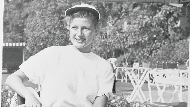 rita hayworth sits on a chair and holds two tennis rackets next to her, she wears a baseball cap, tshirt and shorts, she smiles and looks left of the camera