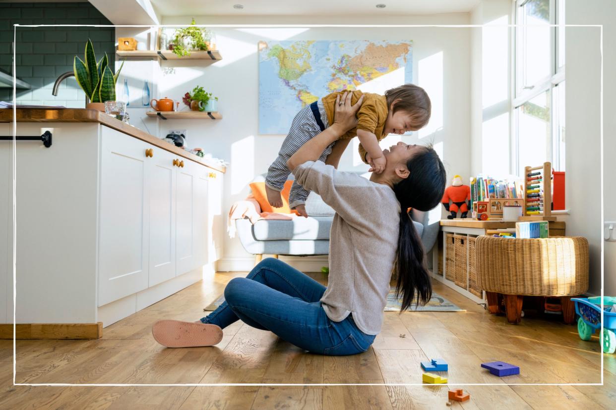  Mother holding her child up in the air child sitting on the floor at home surrounded by toys. 