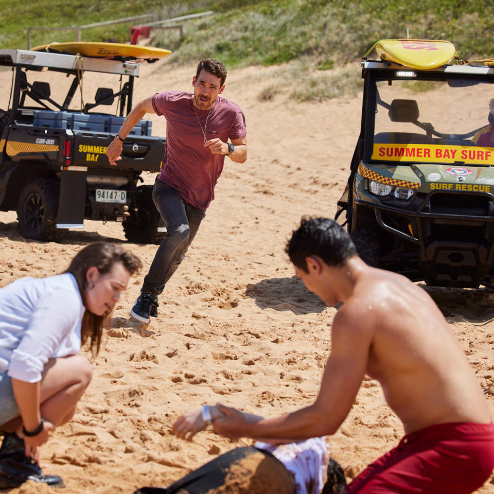 On Palm Beach, Summer Bay, Luke Van Os as character Xander is running on the beach to help rescue Millie. Bella and Nikau are tending to Millie after she was rescued from the waves.