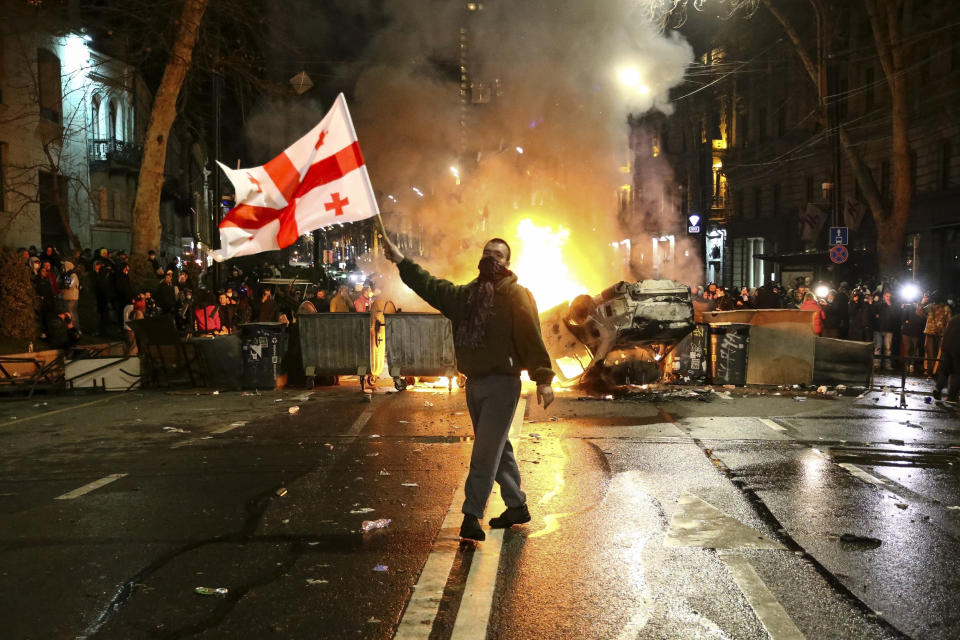 A man waves a Georgian flag in front of a burning barricade as other protesters stand behind not far from the parliament building in Tbilisi on March 9, 2023.  (Zurab Tsertsvadze / AP)