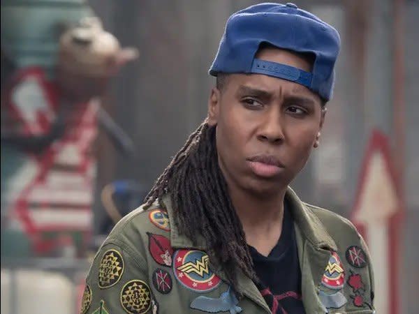 lena waithe as aech/helen from ready player one wears a blue hat and green jacket with patches on it