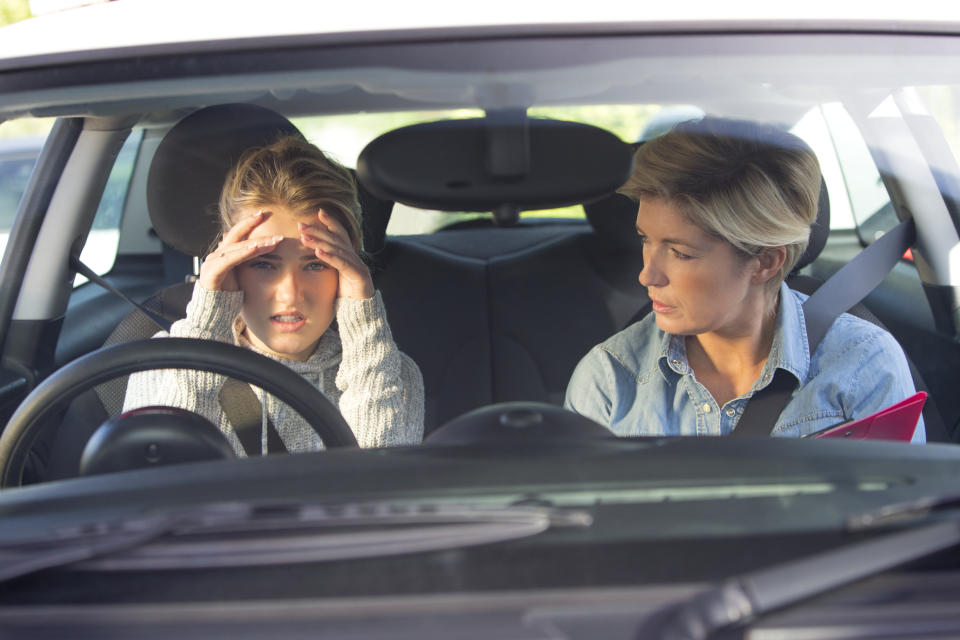 A stressed teenage girl sits in the driver's seat with her hands on her head, while an older woman, possibly her mother or driving instructor, looks at her with concern from the passenger seat