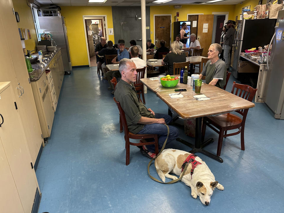 Ron Adams, left foreground with service dog, sits down to dinner across from Scott Snyder at an emergency shelter for homeless men run by St. Elizabeth Shelters in Santa Fe, N.M., on Monday, May 22, 2023. A new tally of the homeless population in New Mexico shows an abrupt jump in the number of people living without permanent housing or with no shelter at all amid surging prices for rent. (AP Photo/Morgan Lee)