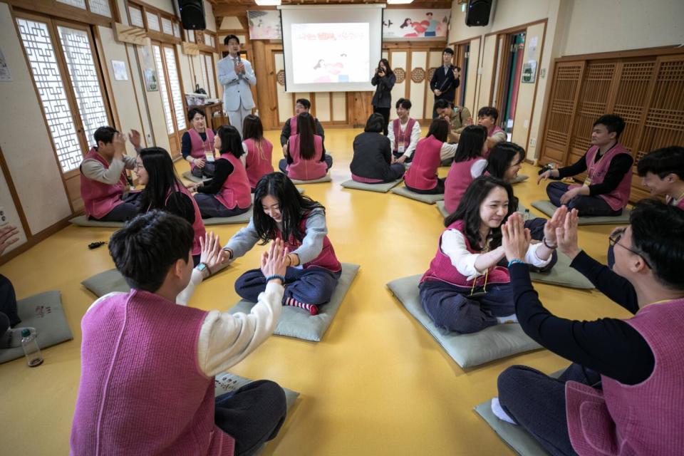 Men and women, in pairs, touch their palms together while seated on mats in a row