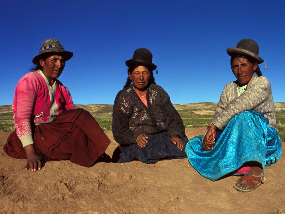 A photo of three women sitting on the ground all wearing bowler hats and colorful skirts.