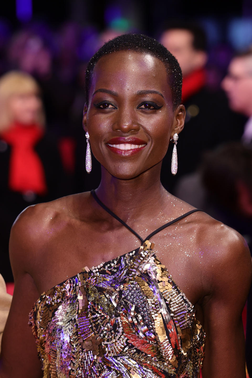 Lupita Nyong'o smiling in a metallic dress with sequins and long, dangling earrings on the red carpet