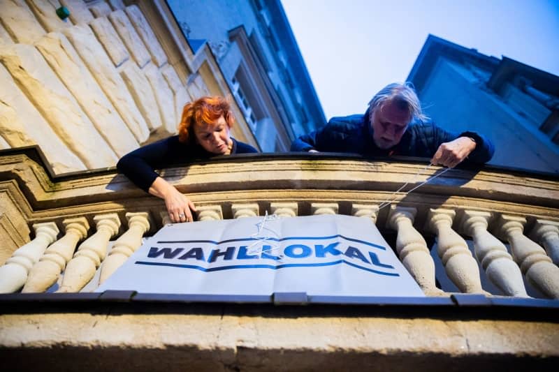 Two poll workers hang a sign at the polling station in the Carl-von-Ossietzky-Gymnasium in Berlin-Pankow before voting begins. Voters in hundreds of Berlin constituencies head to the polls for a partial re-run of the 2021 federal election, after numerous mishaps the first time round. Christoph Soeder/dpa
