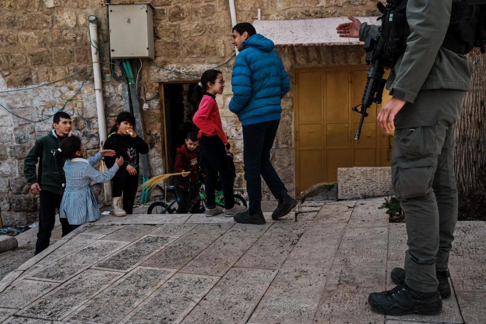 A soldier pictured from below the shoulders signals to several children playing outside, one with a bike