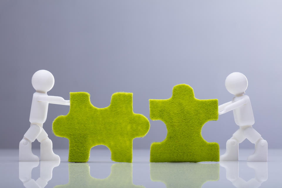 Two miniature human figurines pushing together two green jigsaw puzzle pieces.
