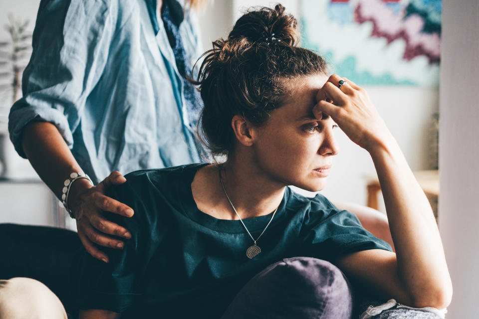 A woman, looking distressed, sits with her hand on her forehead as another person stands beside her, offering comfort with a hand on her shoulder