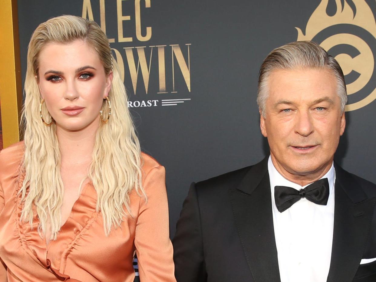 ireland baldwin and alec baldwin at the comedy central roast of alec baldwin. ireland is a young woman with wavy blonde hair, wearing a satin orange dress. she's holding her father's hand. alec baldwin is wearing a black tuxedo with his silver hair slicked back.