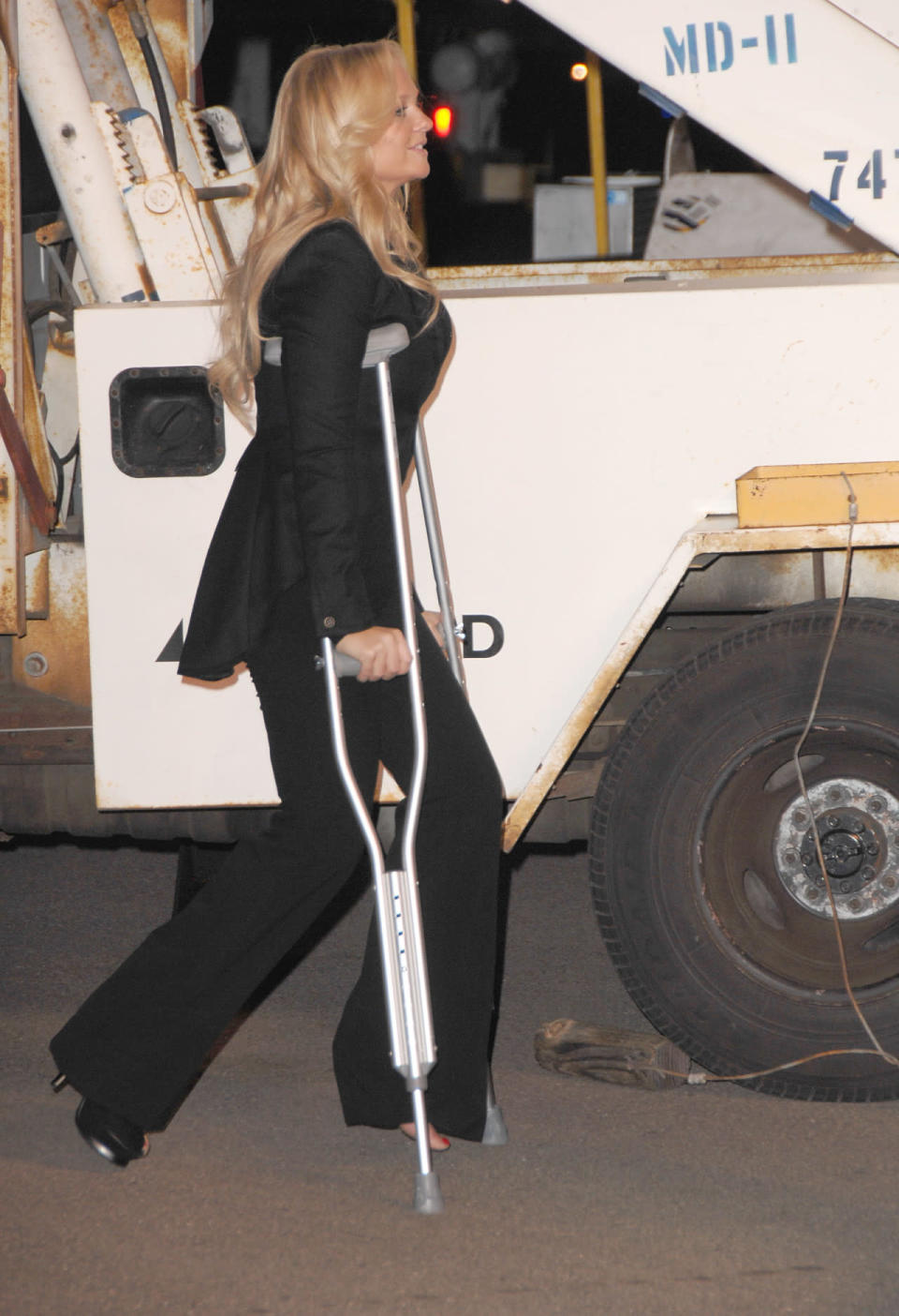 Emma Bunton in a black pantsuit, a single heel, and crutches.