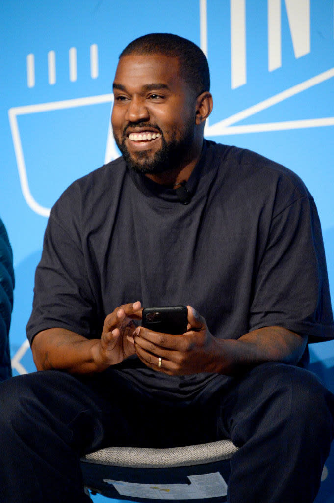 dressed in a casual t-shirt and pants, Kanye sits on stage smiling with his phone in his hands