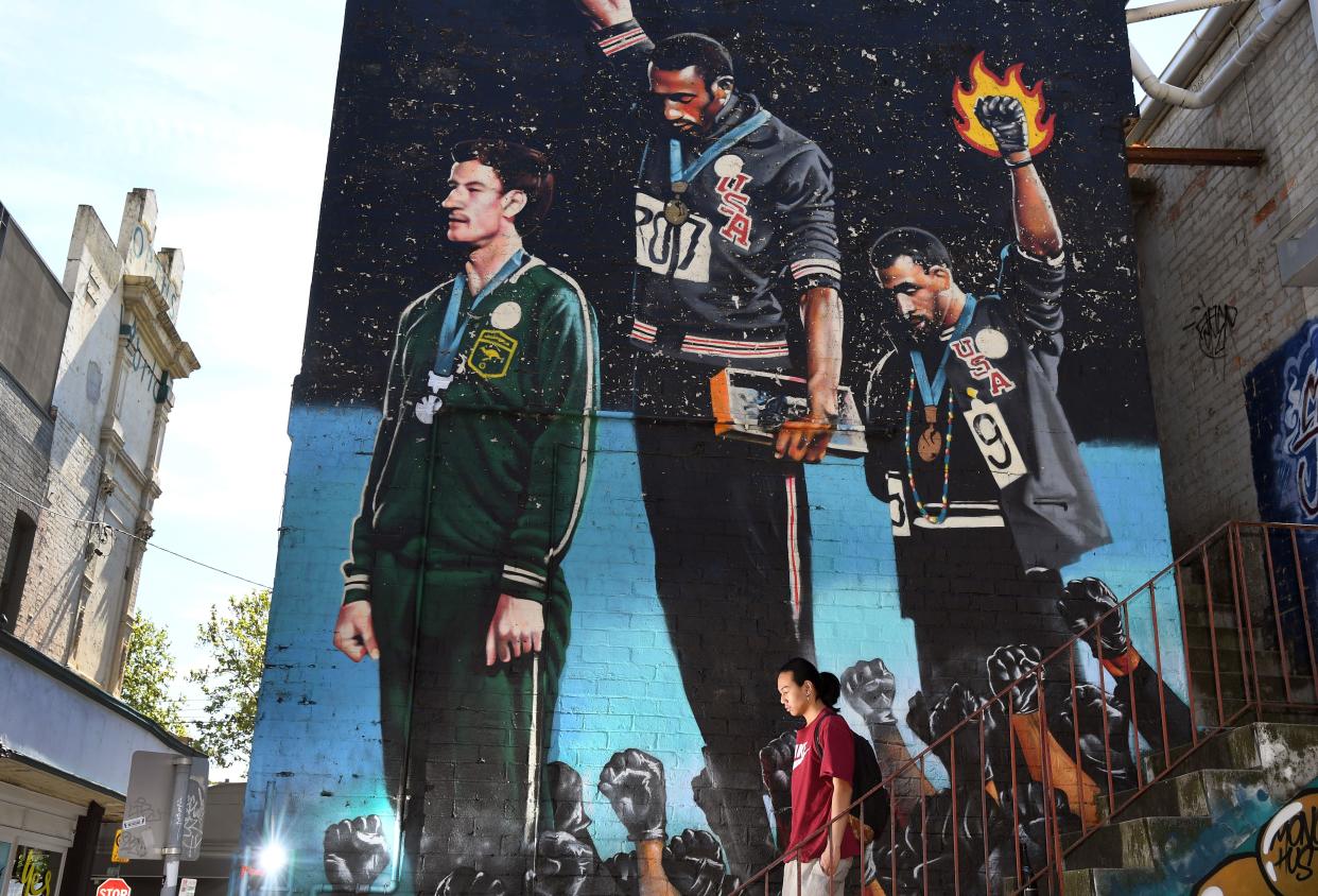 A photo taken in Melbourne on October 8, 2018, shows a man walking past a giant mural of Australian runner Peter Norman with US sprinters Tommie Smith and John Carlos. - When a principled Peter Norman stood on the podium alongside two Americans in their famous Black Power salute at the 1968 Mexico Olympics, his place in the history books was sealed. As sprinters Tommie Smith and John Carlos raised their black-gloved fists in silence, their heads bowed in protest calling for racial equality in the United States, Norman made a choice that lived with him for the rest of his life. (Photo by William WEST / AFP)        (Photo credit should read WILLIAM WEST/AFP via Getty Images)