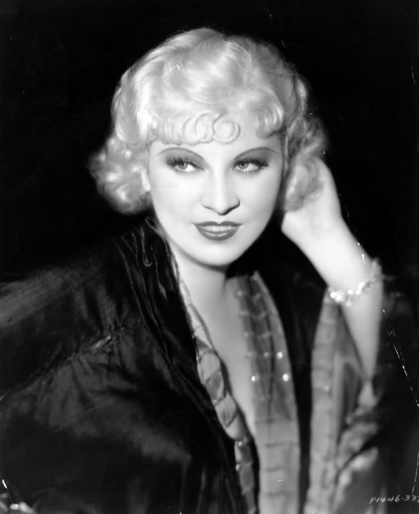 Mae West poses in a glamorous old Hollywood portrait wearing a satin dress with curled hair, a bracelet on her wrist, and a confident expression